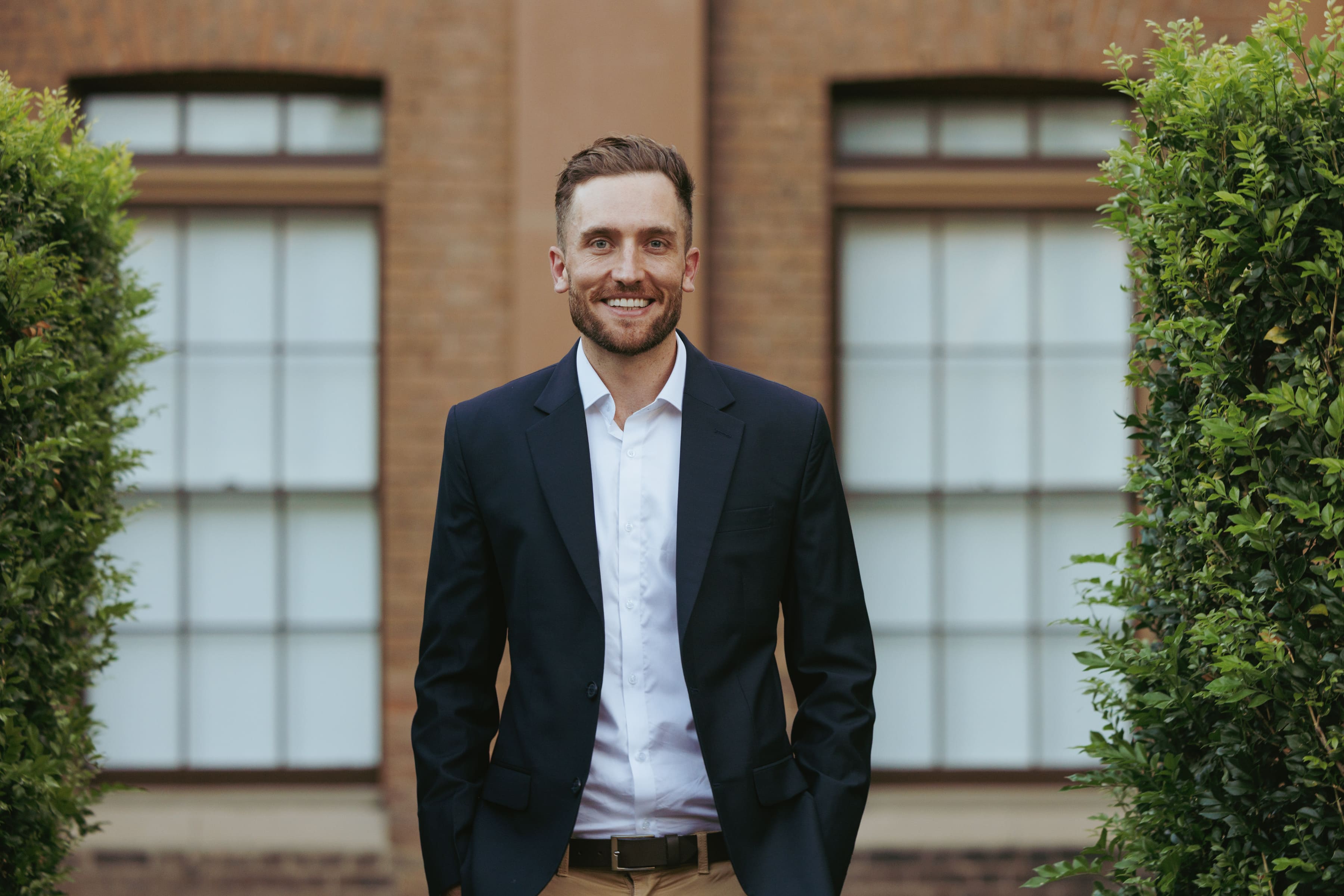 Full-length photo of Andy Crebar standing in a courtyard between hedges, wearing a suit