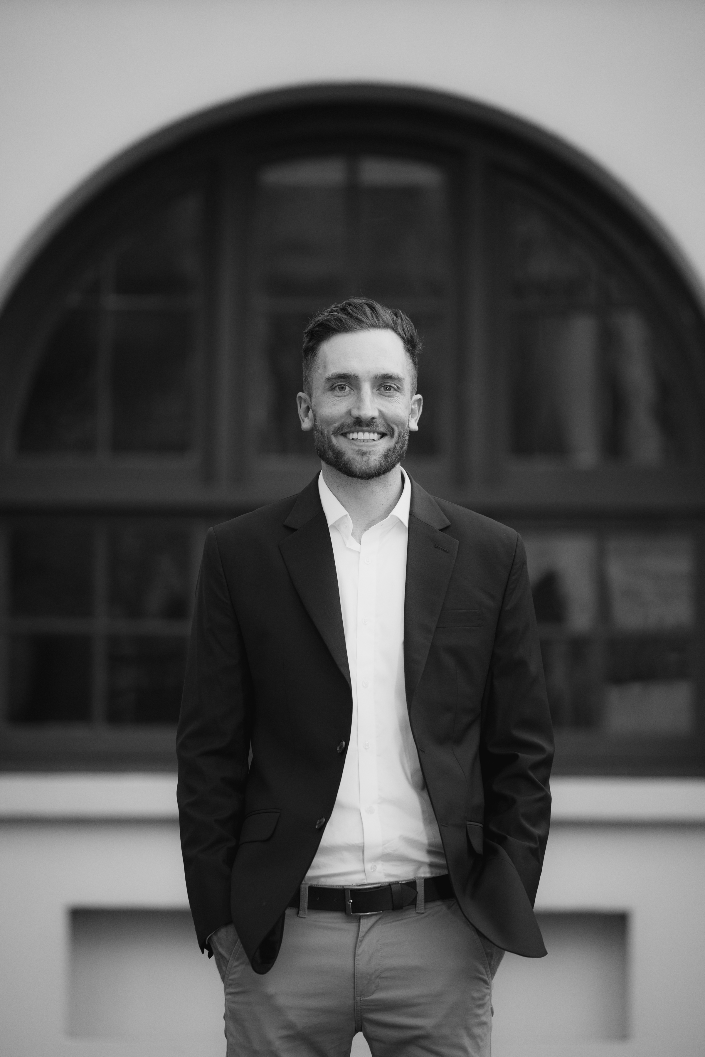 Black and white portrait of Andy Crebar wearing a formal suit in front of an arched window