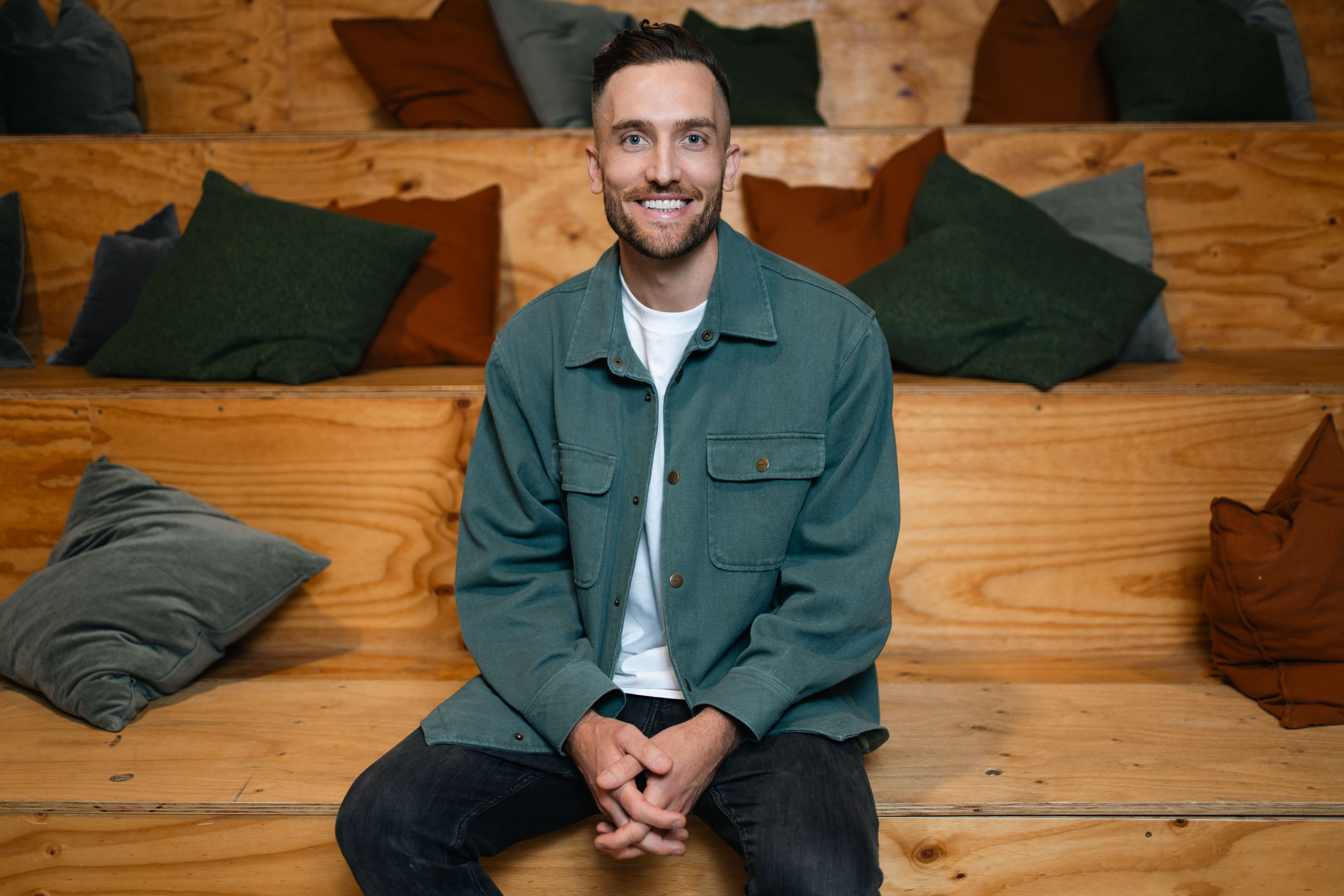 Andy Crebar sitting on wooden tiered seating in a coworking space, wearing a green jacket